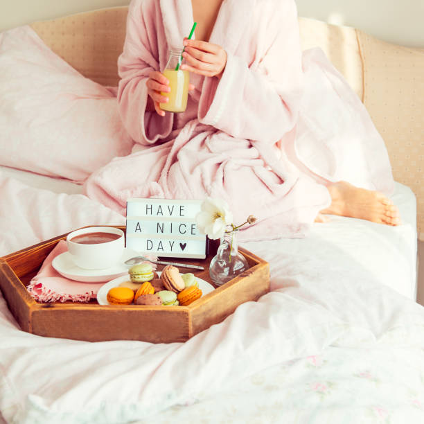 istock Good morning concept. Breakfast in bed with Have a nice day text on lighted box, coffee and macaroons on tray and woman in bathrobe drinking juice. Hospitality, care, service. Square. Copy space. 1130942958