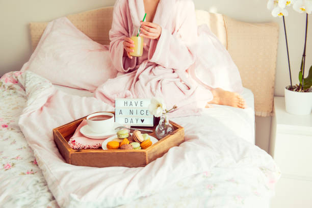 istock Good morning concept. Breakfast in bed with Have a nice day text on lighted box, coffee and macaroons on tray and woman in bathrobe drinking juice. Hospitality, care, service. Square. Copy space. 1130942961