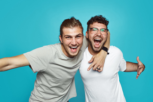 Two friends in casual wear standing and laughing together. Best friends enjoying isolated over blue background. Two men having fun isolated over blue wall.