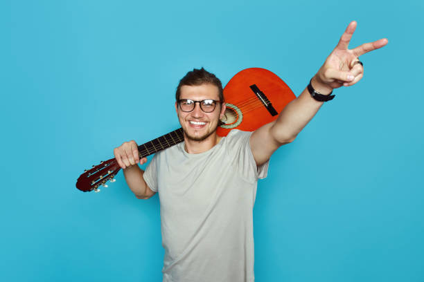 beau garçon de hipster avec la guitare acoustique sur l'épaule souriant et montrant le geste de paix tout en restant sur le fond bleu vif - isolated on white studio shot guitar young men photos et images de collection