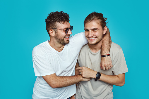 Image of ttwo cheerful handsome guys embracing each other, wearing casual clothes, isolated over blue studio background