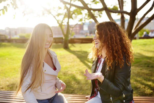 istock Two beautiful young women talking while sitting on a bench at sunny park.Communication and gossip. 1130959213