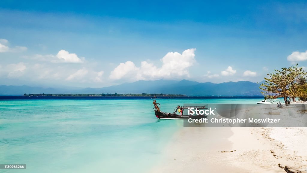 Gili Island - Boat to the Paradice A boat parked next to the shore on Gili Air, Lombok, Indonesia. Beautiful and clear water. In the back visible Mount Rinjani. Some trees on the shore, few clouds on the sky. Bali Stock Photo