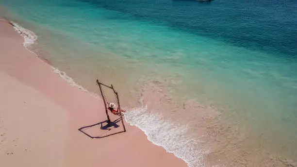 A girl swinging on the beach swing on Pink Beach, Lombok, Indonesia. Captured from above with a drone. The water changes colors from turquoise to navy blue. beach has a nice coral color.