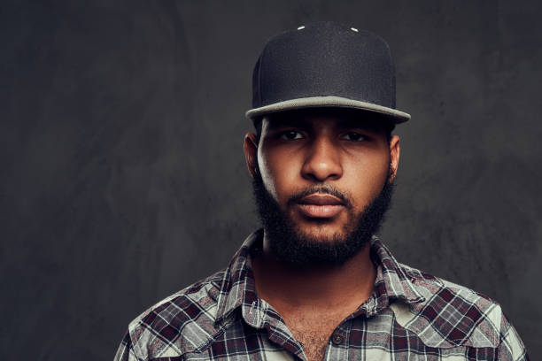 Close-up portrait of African-American guy wearing a checkered shirt and cap standing next to a dark textured background.