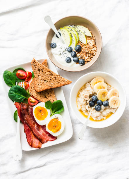 Varied sweet and savory breakfast - bacon with egg and vegetables, spinach salad, yogurt with granola and fruit, vegetarian oatmeal on a light background, top view