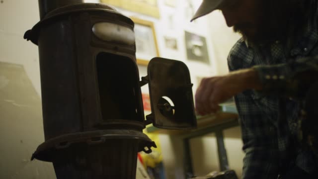 A Middle-Aged Caucasian Man Picks up Wood Kindling and Places It in a Wood Burning Stove in a Workshop