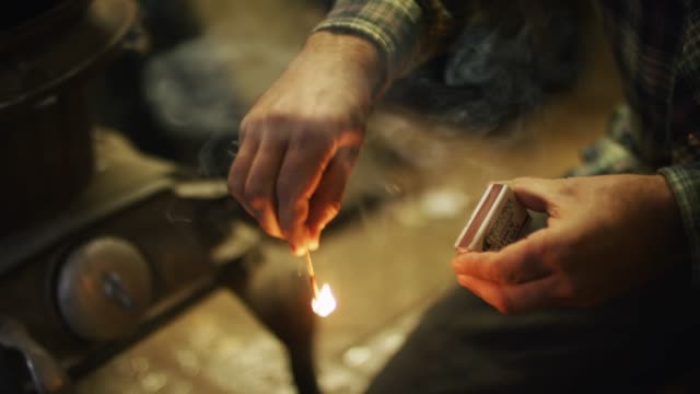 A Man's Hands Strike a Match and Throws the Lighted Match into a Wood Burning Stove in a Workshop