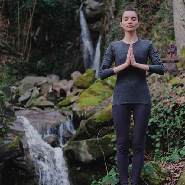 istock Young slim woman practicing yoga outdoors in moss forest on background of waterfall. Unity with nature concept. Girl standing with prayer hands 1130986767