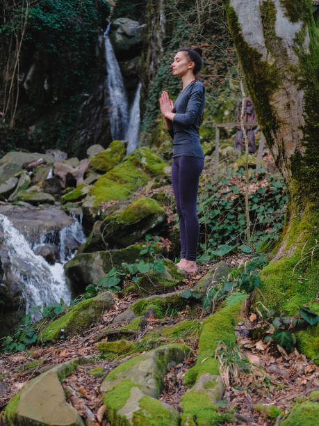 istock Young slim woman practicing yoga outdoors in moss forest on background of waterfall. Unity with nature concept. Girl standing with prayer hands 1130986769
