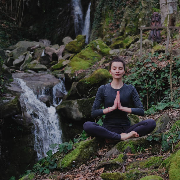 istock Young slim woman practicing yoga outdoors in moss forest on background of waterfall. Unity with nature concept. Girl meditates sitting 1130986770