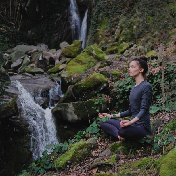 istock Young slim woman practicing yoga outdoors in moss forest on background of waterfall. Unity with nature concept. Girl meditates sitting 1130986773