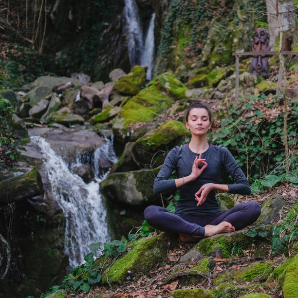 istock Young slim woman practicing yoga outdoors in moss forest on background of waterfall. Unity with nature concept. Girl meditates sitting 1130986774