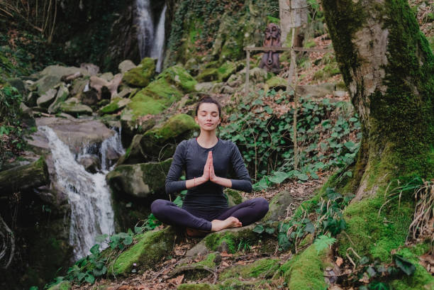 istock Young slim woman practicing yoga outdoors in moss forest on background of waterfall. Unity with nature concept. Girl meditates sitting 1130986779