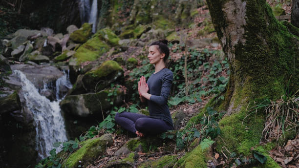 istock Young slim woman practicing yoga outdoors in moss forest on background of waterfall. Unity with nature concept. Girl meditates sitting 1130986783