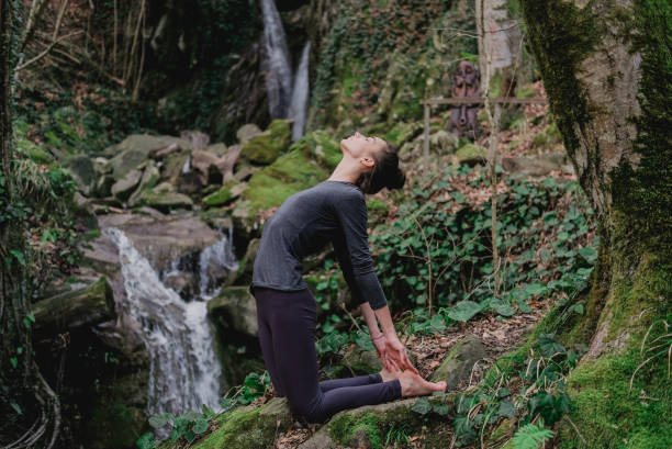 istock Young slim woman practicing yoga outdoors in moss forest on background of waterfall. Unity with nature concept. Girl in camel position 1130986796