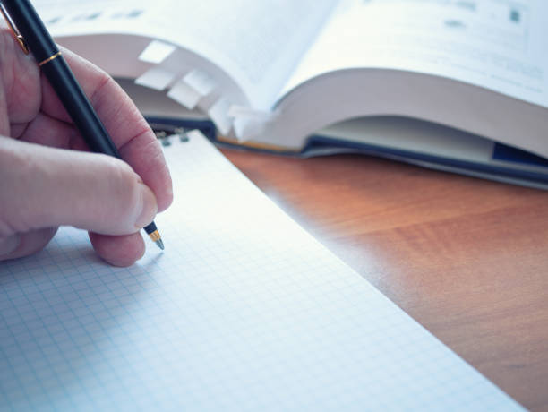 istock left handed student with pen preparing for exams, writing notes at textbook on wooden desk. opened encyclopedia dictionary with bookmarks on background out of focus. closeup image 1130992698