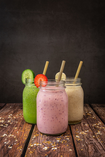 A Strawberry, blueberry and banana smoothie.  A cucumber, spinach, Lime and avocado smoothie.  A Banana and pineapple smoothie.  All with flax seed, chia seed and oat smoothie in drinking jars photographed on a wooden table against a dark background.  Also pictured is bamboo eco straws.