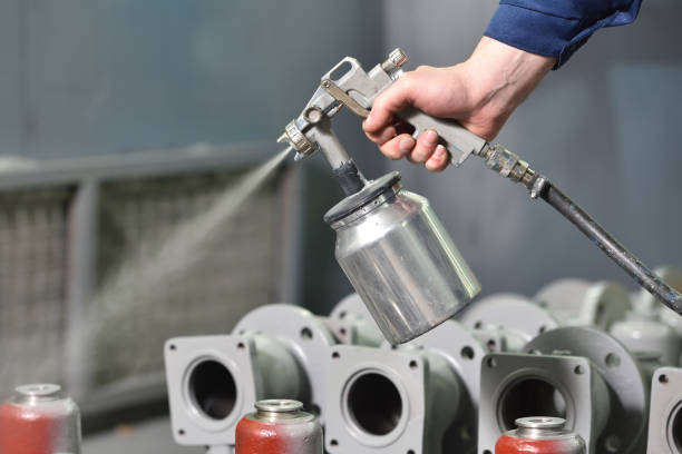 A worker at a factory in a special room paints parts of the valves from the spray gun. Paint valves in gray color, hand and spray gun close up stock photo