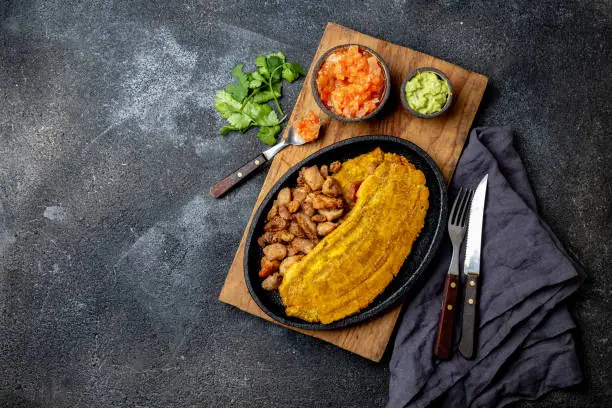 Photo of COLOMBIAN CARIBBEAN CENTRAL AMERICAN FOOD. Patacon or toston, fried and flattened whole green plantain banana on white plate with tomato sauce and chicharron Black background, top view