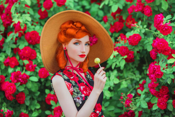 винтажная девушка с красными губами в платье с принтом роз с красными конфетами на летнем огороде. рыжая модель в летней шляпе на фоне кусто - women lollipop old fashioned red hair стоковые фото и изображения