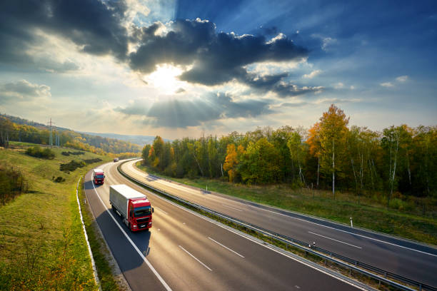 istock Three red trucks driving on the asphalt highway between deciduous forest in autumn colors under the rays of the sunset and dramatic clouds. View from above. 1131044100