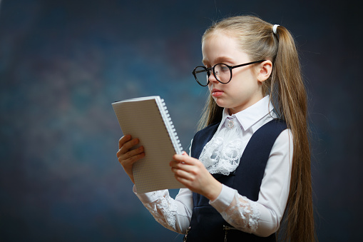 Schoolgirl Wear Glasses Uniform Look at Notebook. Camera Shot on Dark Background. Pensive Little Child with Writing Pad. Clever Elementary School Scholar Study, get Knowledge, do Homework