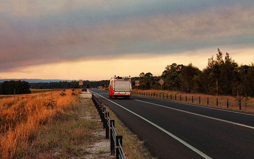 A rural fire service, fire and rescue vehicle responds urgently to a large out of control bushfire in the mountains.  Smoke and sun casts a red glow across the landscape. Thick smoke billowing overhead