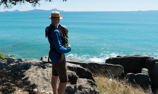 Young father with toddler in a carry pack enjoys a day hike on a coastal walkway with stunning views of the ocean
