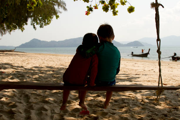 Two brothers seat on a swing in a Thailand beach, back point of view with  the sea as background stock photo