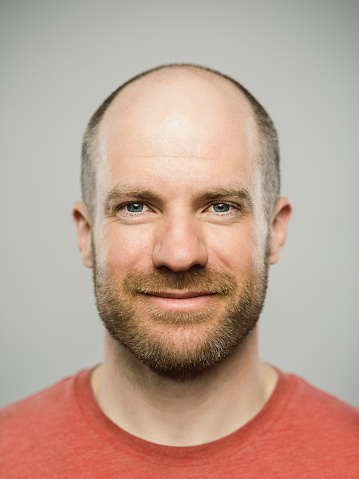 Close up portrait of mature adult adult caucasian man with happy expression looking at camera against white gray background. Vertical shot of canadian real people smiling in studio with short balding read hair and blue eyes. Photography from a DSLR camera. Sharp focus on eyes.