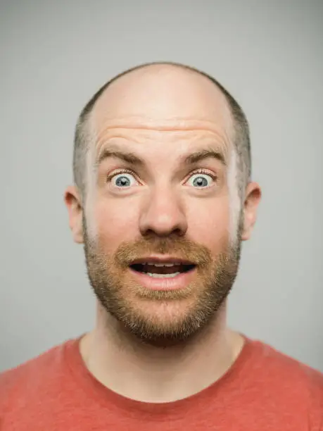 Close up portrait of mature adult adult caucasian man with surprised expression looking at camera against white gray background. Vertical shot of canadian real people with awe reaction in studio with short balding read hair and blue eyes. Photography from a DSLR camera. Sharp focus on eyes.