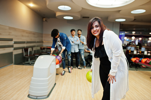 South asian woman standing at bowling alley with ball on hands. Girl is preparing for a throw.