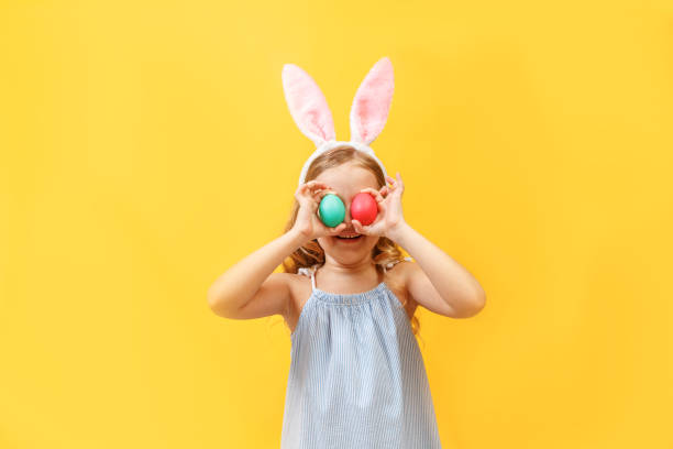 istock Cheerful little child girl with bunny ears with two Easter eggs on a colored background. 1131095838
