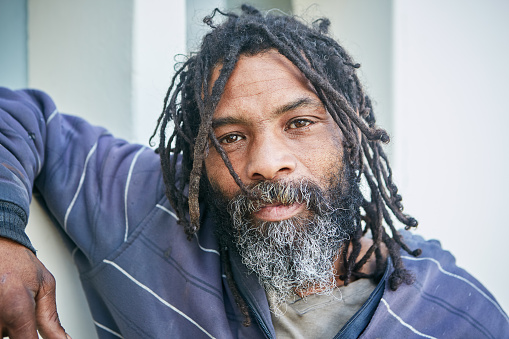 A homeless man with dreadlocks leans on a wall and looks at camera with a thoughtful but resigned expression,