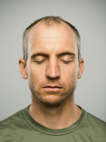 Close up portrait of mid adult caucasian man with blank expression and eyes closed against gray white background. Vertical shot of american real people resting in studio with short balding hair and blue eyes. Photography from a DSLR camera. Sharp focus on eyes.