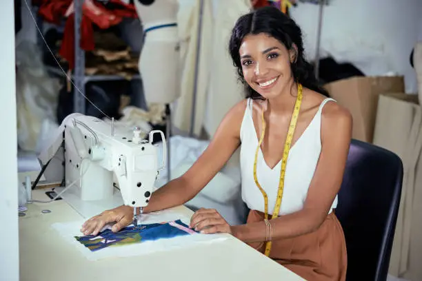Beautiful smiling young seamstress working on sewing machine in workshop