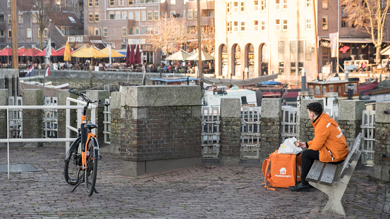 Rotterdam The Netherlands - February 14, 2019: Food delivery guy of Thuisbezorgd.nl (Delivered at home) having a break nearby the Old Harbor at sunset