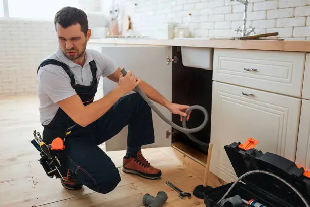 Photo of Young man hold hose in hand. Bad smell. Stinks. Handyman suffer from it. Opened toolbox and piece of pipe on floor.