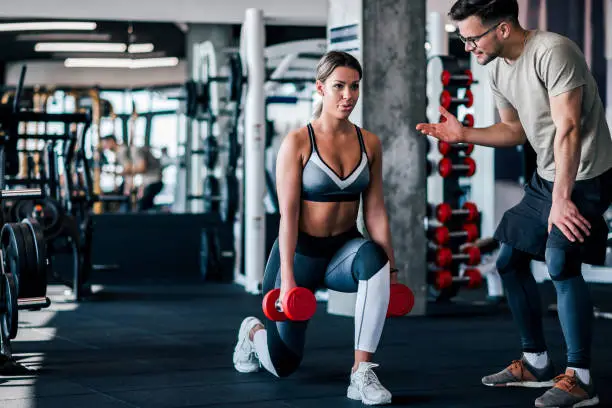 Photo of Young muscular woman doing weighted lunge with dumbbells, with personal trainer motivating her.