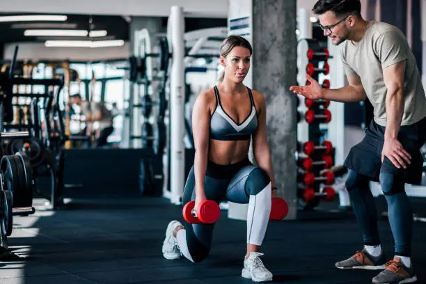 Young muscular woman doing weighted lunge with dumbbells, with personal trainer motivating her.