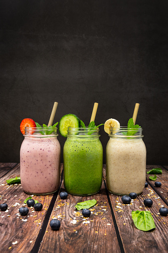 A Strawberry, blueberry and banana smoothie.  A cucumber, spinach, Lime and avocado smoothie.  A Banana and pineapple smoothie.  All with flax seed, chia seed and oat smoothie in drinking jars photographed on a wooden table against a dark background.  Also pictured is bamboo eco straws and sprigs of mint.