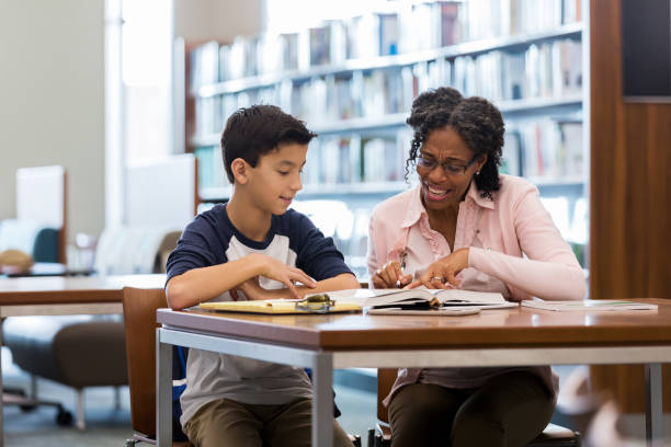 niño de la edad de la escuela media lee con mentor en biblioteca - maestro particular fotografías e imágenes de stock