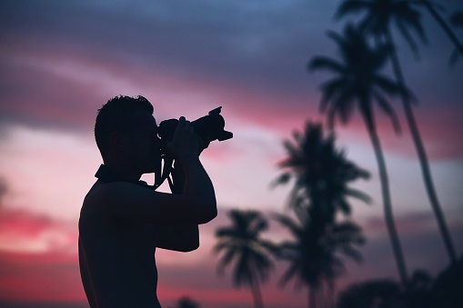 Silhouette of the young photographer with camera against palm tree at colorful sunset.
