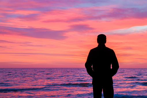 Young adult man in warm black overcoat standing alone and staring at small waves of sea and colorful sunset light in sky. Peaceful atmosphere in winter evening. Back view.