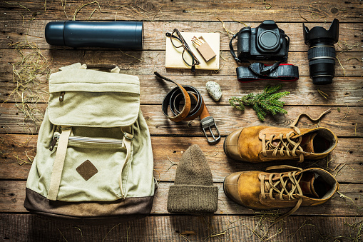 Traveling - packing (preparing) for adventure trip concept. Backpack, boots, hat, belt, thermos and camera on wooden background captured from above (flat lay).