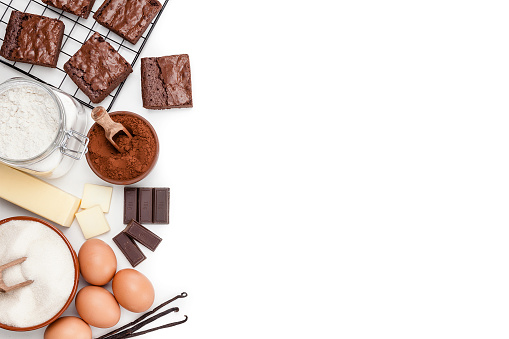 Top angle view of ingredients for preparing homemade chocolate brownies arranged at the left of an horizontal frame leaving useful copy space for text and/or logo. Ingredients included in the composition are butter, sugar, flour, eggs, chocolate bars and salt. Various baked brownies are at the top placed in a cooling rack. Predominant colors are brown and white. High key DSRL studio photo taken with Canon EOS 5D Mk II and Canon EF 70-200mm f/2.8L IS II USM Telephoto Zoom Lens