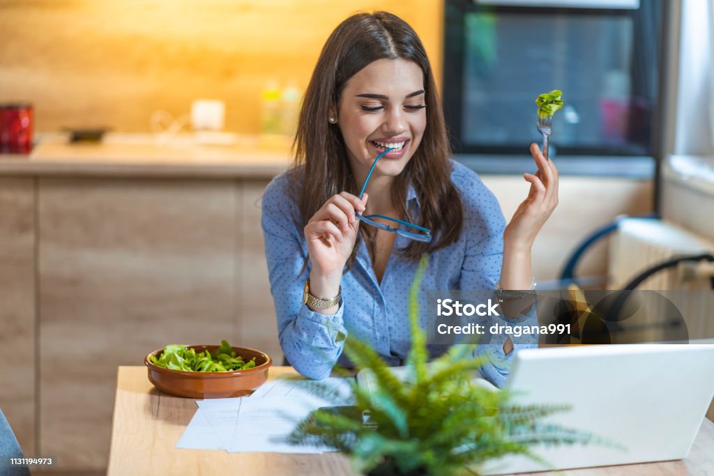 Young business woman eating salad at home Young business woman eating salad at home, having healthy lunch at workplace. Portrait of pretty young woman eating salad and working at home. Eating Stock Photo