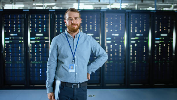 istock Bearded IT Specialist in Glasses is Standing in Data Center Next to Server Racks, Smiling to the Camera and Holding Laptop. Running Diagnostics or Doing Maintenance Work. 1131198054