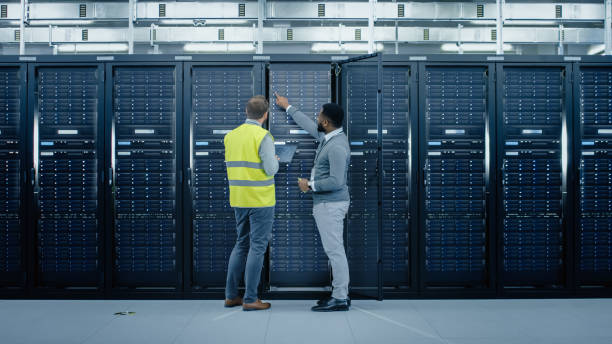 istock Bearded IT Engineer in Glasses and High Visibility Vest with a Laptop Computer and Black Technician Colleague Talking in Data Center while Standing Next to Server Racks. Running Diagnostics or Maintenance. 1131198057